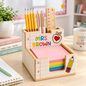 Puede incluir: Un organizador de escritorio de madera con diseño de papel de cuaderno, con el texto MRS. BROWN, un dibujo de manzana roja y un lápiz. Contiene cuatro lápices amarillos, dos bolígrafos rosas, una regla de madera y notas adhesivas de colores.