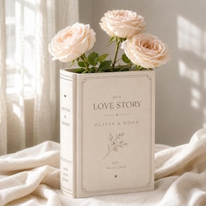 May include: A beige book-shaped vase featuring the text OUR LOVE STORY, OLIVIA & NOAH, and EST. 04.12.2025, decorated with a floral line drawing and three pale pink roses emerging from the top, with the names OLIVIA & NOAH printed on the spine.