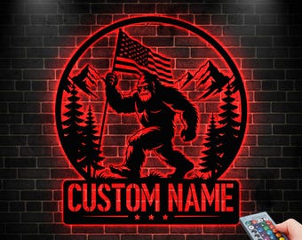 May include: A red-lit metal sign featuring a silhouette of Bigfoot holding an American flag, set within a circular frame with mountains and trees. The sign includes the text "CUSTOM NAME" in a rectangular panel below. The background is a dark brick wall.