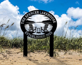 May include: Black metal lake house sign with the text "The Spencer Lakehouse Established 2025". The sign features a lake scene with two Adirondack chairs and a small table. The sign is on a beach with grass and a blue sky.