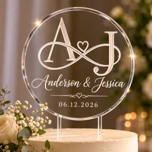 May include: A circular clear acrylic cake topper featuring the engraved letters A and J connected by an infinity symbol with a small heart. Below, the names Anderson & Jessica are printed with a date of 06.12.2026 and a small heart icon.