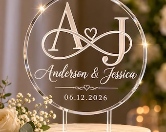 May include: A circular clear acrylic cake topper featuring the engraved letters A and J connected by an infinity symbol with a small heart. Below, the names Anderson & Jessica are printed with a date of 06.12.2026 and a small heart icon.