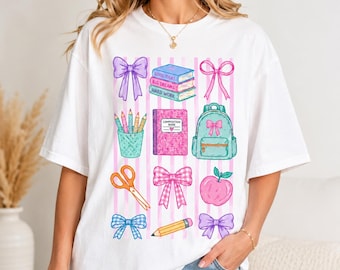 May include: A white short-sleeve t-shirt featuring a colorful graphic print of school supplies including a stack of books labeled Good Ideas, Big Dreams, Hard Work, a pencil cup, a backpack, scissors, an apple, a notebook, and various bows.