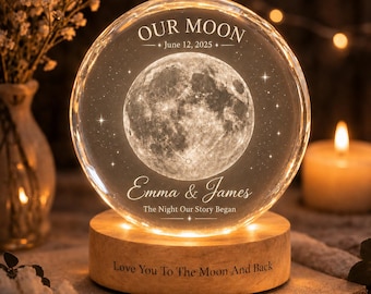 May include: A circular glass orb lamp featuring a laser-etched moon and stars. The text reads "OUR MOON", "June 12, 2025", "Emma & James", and "The Night Our Story Began". The wooden base is engraved with "Love You To The Moon And Back".
