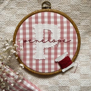 May include: A circular wooden embroidery hoop containing pink and white gingham fabric. A large white fabric letter P is appliqued in the centre, with the name penelope embroidered in dark red cursive thread. A small spool of red thread sits on the fabric.