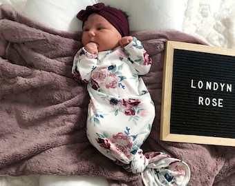 Könnte beinhalten: Ein Baby trägt ein weißes, geknotetes Kleid mit Blumendruck und ein burgunderfarbenes Stirnband. Das Kleid hat ein burgunderfarbenes, rosa und blaues Blumenmuster. Das Baby liegt auf einer lila Decke mit einem schwarzen Buchstabenbrett, auf dem "LONDYN ROSE" steht.