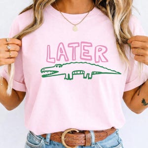 Könnte beinhalten: Ein hellrosa T-Shirt mit dem Wort LATER in fetter rosa Schrift über einer grünen Strichzeichnung eines Alligators. Das Shirt hat einen Rundhalsausschnitt und kurze Ärmel und zeigt ein einfaches grafisches Design auf der Vorderseite.