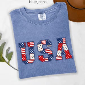 Könnte beinhalten: Hellblaues T-Shirt mit dem Wort "USA" in einem patriotischen Design. Die Buchstaben sind mit rot-weißen Streifen und blauen Sternen gefüllt. Das Shirt ist gefaltet und hat ein Etikett mit der Aufschrift "Comfort Colors".