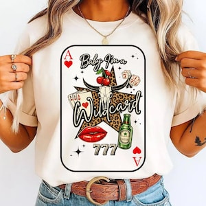 Pode incluir: Camiseta creme com um design de carta de baralho. O design inclui uma estrela com estampa de leopardo, um crânio com cerejas, dados e as palavras "Baby I'm a Wildcard". Um lábio vermelho, uma garrafa de cerveja e o número 777 também estão presentes.