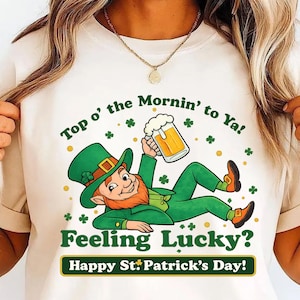 Könnte beinhalten: Cremefarbenes T-Shirt mit einem Cartoon-Kobold in grüner Kleidung, der ein Bier hält. Der Text "Top o' the Mornin' to Ya!" und "Feeling Lucky? Happy St. Patrick's Day!" sind ebenfalls zu sehen.
