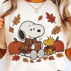 Könnte beinhalten: Ein cremefarbenes T-Shirt mit einem Grafikdruck von Snoopy und Woodstock, die zwischen orangefarbenen Kürbissen und herbstlichen Blättern sitzen. Snoopy trägt einen mehrfarbigen gestreiften Schal und hält eine Tasse, während Woodstock eine kleinere Tasse hält.