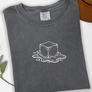 Könnte beinhalten: Dunkelgraues T-Shirt mit einer weißen Umrissgrafik eines schmelzenden Eiswürfels. Das Shirt ist gefaltet, mit einem Etikett am Halsausschnitt. Ein brauner Hut ist teilweise im Hintergrund zu sehen.