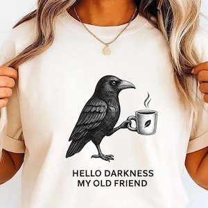 Könnte beinhalten: Cremefarbenes T-Shirt mit einer Illustration eines schwarzen Raben, der eine Kaffeetasse hält. Der Text "HELLO DARKNESS MY OLD FRIEND" ist darunter gedruckt. Das Design ist eine grafische Illustration.