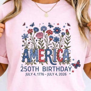 Könnte beinhalten: Hellrosa T-Shirt mit dem Wort "AMERICA" in Blau und Rot, verziert mit einem Blumendesign und Schmetterlingen. Der Text "250TH BIRTHDAY JULY 4, 1776 - JULY 4, 2026" steht darunter.