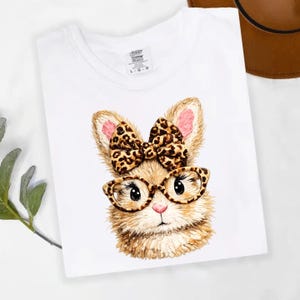 Könnte beinhalten: Weißes T-Shirt mit einem Cartoon-Hasen, der eine Brille und eine Schleife trägt. Der Hase hat braunes Fell, rosa Ohren und eine rosa Nase. Die Schleife und die Brillengestelle haben ein Leopardenmuster. Das T-Shirt hat einen Rundhalsausschnitt.