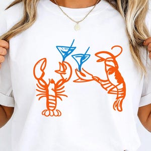 Könnte beinhalten: Weißes T-Shirt mit einem orangefarbenen Hummer-Design, das ein blaues Cocktailglas hält. Ein zweiter Hummer hält ein zweites blaues Cocktailglas. Das Design ist eine lustige, skurrile Illustration, ideal für eine Sommer- oder Strand-Themen-Garderobe.