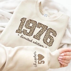 Può includere: Felpa color crema con l'anno "1976" in un design a stampa leopardata, con le parole "limited edition" sotto. La manica presenta il testo "chapter 50" con un cuore leopardato.