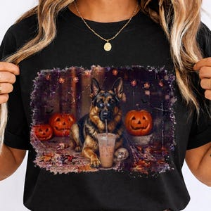 Könnte beinhalten: Ein schwarzes T-Shirt mit einem Grafikdruck eines Deutschen Schäferhundes, der eine Sonnenbrille trägt und aus einem durchsichtigen Plastikbecher mit Strohhalm trinkt. Der Hund ist von zwei geschnitzten Kürbissen und Herbstlaub umgeben.