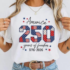 Könnte beinhalten: Weißes T-Shirt mit dem Text "America 250" in Rot und Blau, mit Sternen. Das Shirt zeigt auch "years of freedom 1776-2026". Das Design ist patriotisch und feiert das 250-jährige Jubiläum.