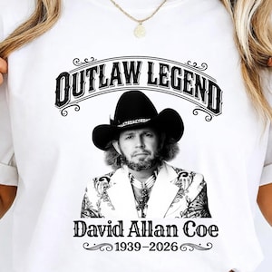 Könnte beinhalten: Weißes T-Shirt mit einem Schwarz-Weiß-Porträt von David Allan Coe, dem Text "OUTLAW LEGEND" darüber und "David Allan Coe 1939-2026" darunter. Das Design enthält dekorative Verzierungen.