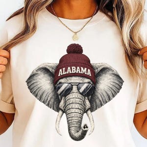 Könnte beinhalten: Cremefarbenes T-Shirt mit einer detaillierten Grafik eines Elefanten, der eine Sonnenbrille und eine weinrote Mütze mit dem Schriftzug "ALABAMA" in weißen Buchstaben trägt. Der Rüssel des Elefanten ist eingerollt. Das Design ist auf dem Shirt zentriert.