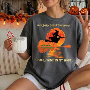 Może przedstawiać: Ciemnoszary t-shirt z grafiką przedstawiającą czarownicę lecącą na miotle na tle dużego pomarańczowego księżyca. Napis głosi ON A DARK DESERT HIGHWAY oraz COOL WIND IN MY HAIR. Wzór zawiera odbicie księżyca i drzew w wodzie.