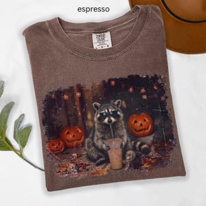 Könnte beinhalten: Ein espressofarbenes T-Shirt mit einer Grafik eines Waschbären, der eine Sonnenbrille trägt und einen Plastikbecher hält. Die Szene zeigt zwei Kürbisse, herbstliche Blätter und eine Lichterkette vor einem dunklen Hintergrund. Label: Comfort Colors.