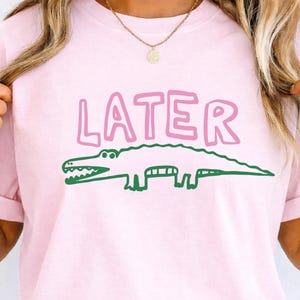 May include: A light pink t-shirt featuring the word LATER printed in large, bubble-style pink letters above a simple, hand-drawn style green alligator illustration. The design is centered on the chest of the garment.