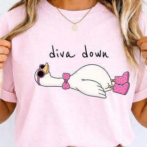 May include: Pink t-shirt featuring a cartoon goose wearing sunglasses, a bow tie, and boots, with the words "diva down" above the goose. The goose is lying down. The shirt is a light pink color.