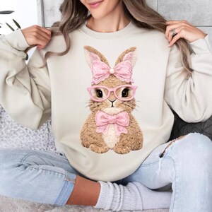 May include: Cream-colored sweatshirt featuring a charming bunny illustration. The bunny is depicted with pink glasses, a bow tie, and a bow on its head. The design is centered on the front of the long-sleeved top, perfect for Easter or spring.