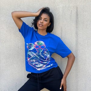 Puede incluir: Una mujer con una camiseta azul con un gráfico de un extraterrestre con gafas de sol y pinchando. También lleva pantalones negros.