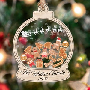May include: Wooden Christmas ornament shaped like a snow globe, featuring gingerbread men and other festive shapes. The ornament has the text "The Walker Family 2025" and a string for hanging. The background is a blurred Christmas tree.