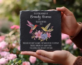 May include: A black slate memorial plaque with the text "In Loving Memory of Brandy Hearne, Beloved Mother 2025." It features colorful hummingbird illustrations and floral accents. The plaque includes a heartfelt message.