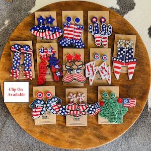May include: A collection of 10 pairs of beaded earrings with patriotic themes. The earrings are displayed on a wooden surface and feature designs such as stars, stripes, boots, cacti, and lightning bolts. The text "Clip On Available" is visible in the image.