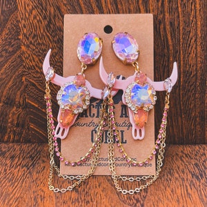 Peut inclure: Boucles d'oreilles en forme de crâne de vache rose et or avec des cristaux irisés et des accents de chaîne rose. Les boucles d'oreilles sont sur un fond brun avec une carte qui dit "Cactus and Coral Country Boutique".