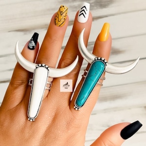 May include: Two silver-toned bull horn rings, one with a white stone and the other with a turquoise stone. The rings are on a hand with painted nails in various colors and designs. The rings have a southwestern or boho style.