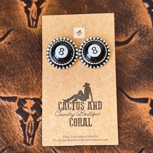 May include: A pair of stud earrings featuring a black and white eight ball design, surrounded by a silver beaded border. The earrings are displayed on a tan card with the text "CACTUS AND COUNTRY BOUTIQUE CORAL".