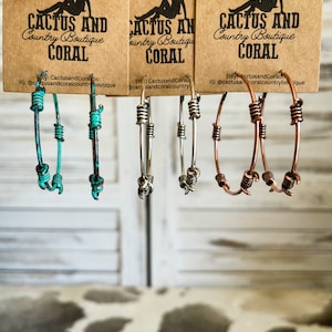 May include: Three pairs of hoop earrings with different colored wire. The earrings are made of turquoise, silver, and copper wire. The earrings are hanging from a brown paper tag with the text "Cactus and Coral Country Boutique" printed on it.