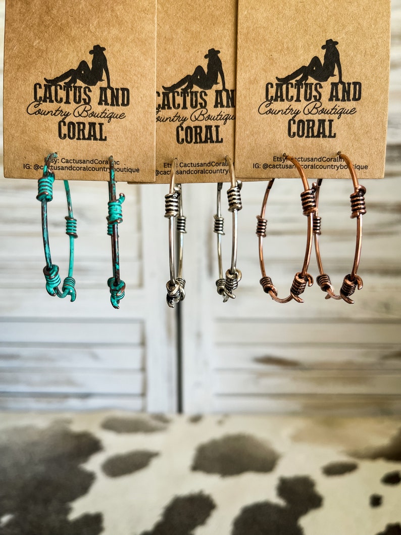 May include: Three pairs of hoop earrings with different colored wire. The earrings are made of turquoise, silver, and copper wire. The earrings are hanging from a brown paper tag with the text "Cactus and Coral Country Boutique" printed on it.
