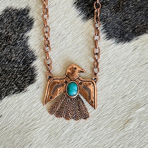 Pode incluir: Colar cor de cobre com um pingente de pássaro estilizado com asas abertas. O pingente tem uma pedra de cor turquesa no centro. O colar é exibido em uma superfície com padrão de pele de vaca.