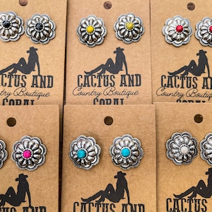 Könnte beinhalten: Ohrstecker mit floralem Design und verschiedenfarbigen Zentren, darunter Schwarz, Gelb, Rot, Rosa, Türkis und Weiß. Die Ohrringe werden auf braunen Karten mit dem Logo "CACTUS AND Country Boutique" präsentiert.