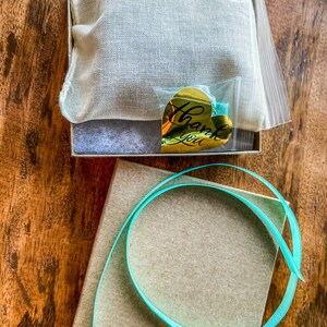 May include: A small brown box with a teal ribbon wrapped around it. The box is sitting on a wooden surface. A smaller box with a white fabric lining and a gold heart-shaped sticker that says "Thank You" is sitting on the wooden surface.