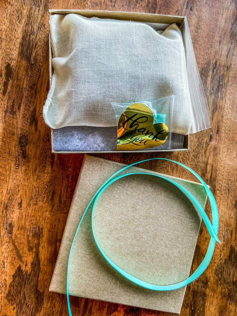 May include: A small brown box with a teal ribbon wrapped around it. The box is sitting on a wooden surface. A smaller box with a white fabric lining and a gold heart-shaped sticker that says "Thank You" is sitting on the wooden surface.