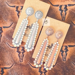 May include: Two pairs of dangle earrings with a silver and copper finish. The earrings feature a large round charm with a floral design and a long, teardrop-shaped charm with iridescent rhinestones.
