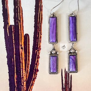 May include: A pair of silver dangle earrings with purple stone accents. The stones are rectangular and have a marbled pattern. The earrings have a star-shaped charm at the top.