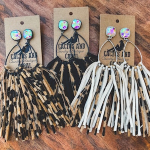 May include: Three pairs of earrings with iridescent square studs and long fringe. The earrings feature a black silhouette of a woman and the text "CACTUS AND Country Boutique CORAL". The fringe is in leopard print, black, and white.