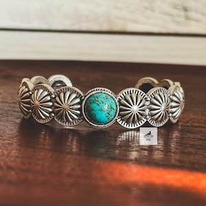 May include: A silver cuff bracelet with a turquoise stone in the center. The bracelet is decorated with a repeating pattern of silver circles with a sunburst design.
