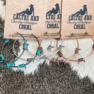 May include: Three pairs of hoop earrings with barbed wire accents. The earrings are in silver, copper, and turquoise colors. The earrings are displayed on a brown and white cowhide background. The text "Cactus and Coral Country Boutique" is visible on the cards behind the earrings.