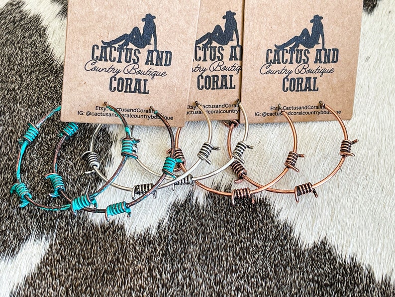 May include: Three pairs of hoop earrings with barbed wire accents. The earrings are in silver, copper, and turquoise colors. The earrings are displayed on a brown and white cowhide background. The text "Cactus and Coral Country Boutique" is visible on the cards behind the earrings.