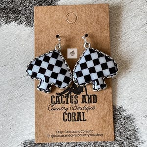 May include: A pair of silver-toned earrings in the shape of a spade, featuring a black and white checkered pattern. The earrings are displayed on a brown card with the text "CACTUS AND Country Boutique CORAL".